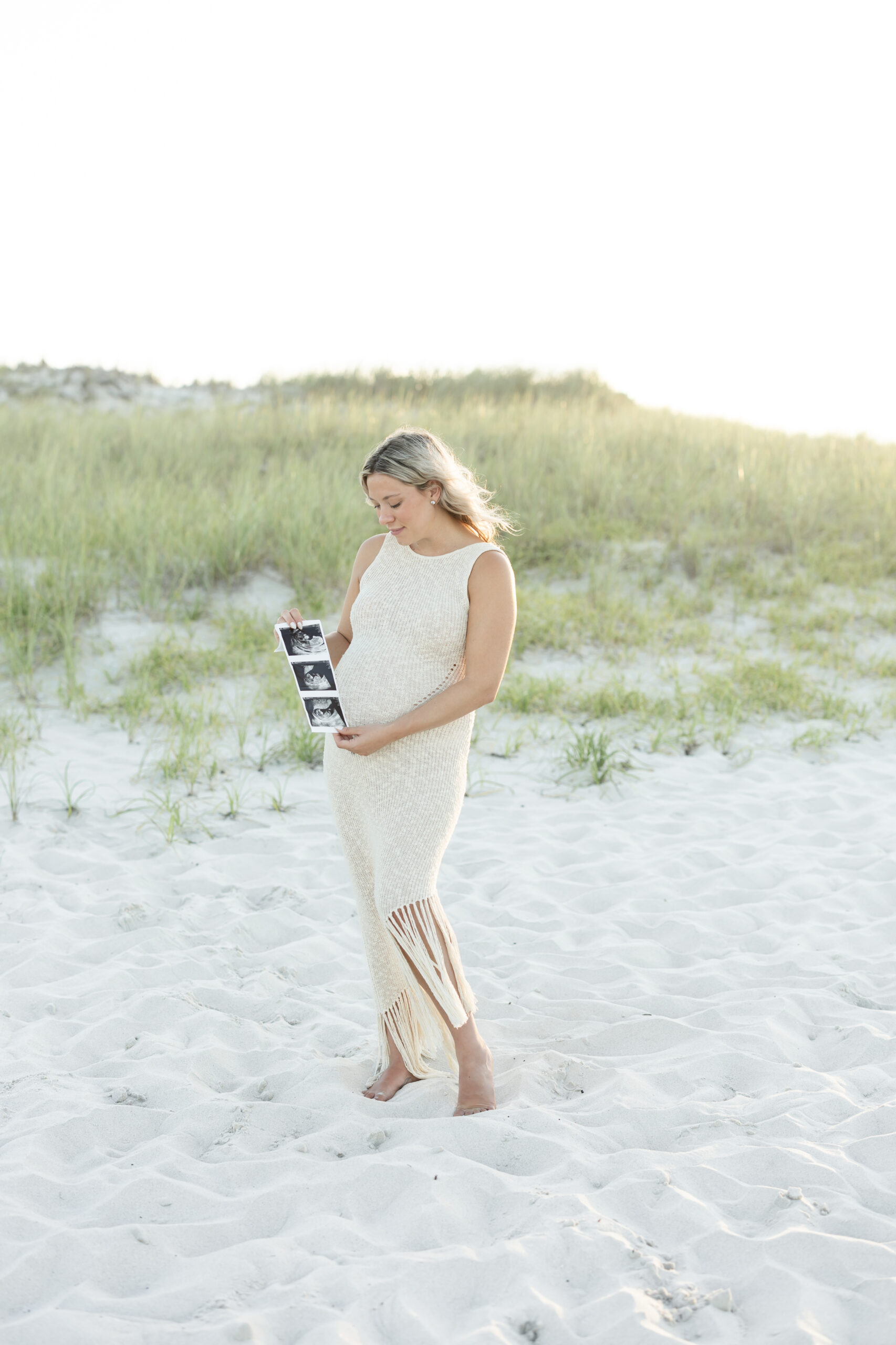 An image of a pregnant mama during her maternity session on Island Beach State Park. She is holding her ultrasound pictures in this image. She was photographed by Natalie Beach Photography, a South Jersey Family Photographer.