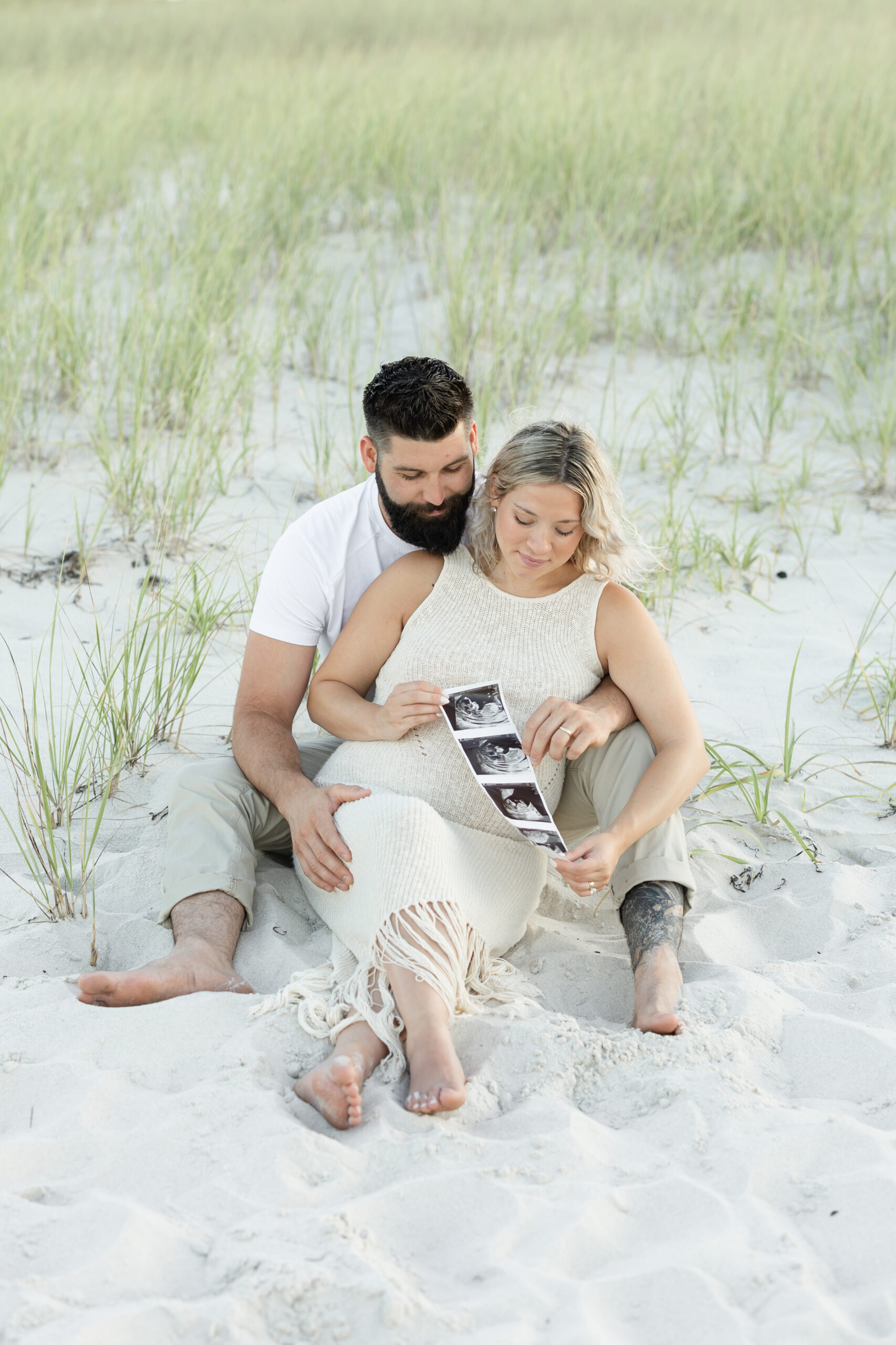 An image of a pregnant mama and her husband during her maternity session on Island Beach State Park. They are sitting among the grassy dunes, holding their ultrasound pictures. They were photographed by Natalie Beach Photography, a South Jersey Family Photographer.